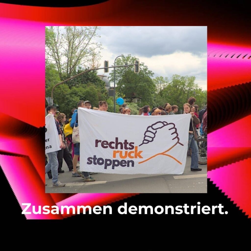 "Bild von Menschen, die ein Banner mit der Aufschrift 'Rechtsruck stoppen' tragen. Sie stehen auf einer Straße und halten das Banner hoch. Hinter ihnen sind Bäume und ein Himmel mit Wolken zu sehen. Das Bild ist von einem roten und pinken Rahmen umgeben."