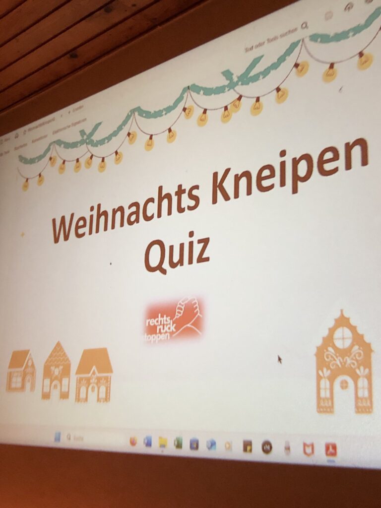 Das Bild zeigt eine Projektion auf einer Leinwand, die einen Titelbildschirm für ein „Weihnachts Kneipen Quiz“ darstellt. Oberhalb des Titels ist eine stilisierte Lichterkette mit grünen Ranken und gelben Glühbirnen abgebildet. Unter dem Titel befinden sich drei kleine, schematisch dargestellte Lebkuchenhäuser, die in Größe und Detaillierung leicht variieren. Rechts unten ist ein größeres, detaillierteres Lebkuchenhaus zu sehen. Im mittleren Bereich des Bildschirms befindet sich das Logo der Organisation „rechts ruck stoppen“, dargestellt durch zwei sich haltenden Hände auf orangem Hintergrund. Der untere Bildrand zeigt einen Ausschnitt der Taskleiste eines Betriebssystems, auf dem verschiedene Programme und Icons erkennbar sind.