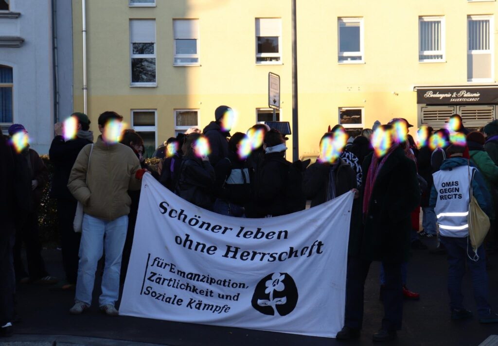 Eine Gruppe von Menschen steht vor einem gelben Gebäude bei einer Demonstration. Sie halten ein weißes Banner mit der Aufschrift "Schöner leben ohne Herrschaft - für Emanzipation, Zärtlichkeit und Soziale Kämpfe". Das Banner zeigt auch eine Blumen-Illustration. Im Hintergrund ist eine Person mit einer Weste zu sehen, auf der "Omas gegen Rechts" steht. Das Gebäude hat mehrere Fenster und eine Bäckerei ist zu erkennen.