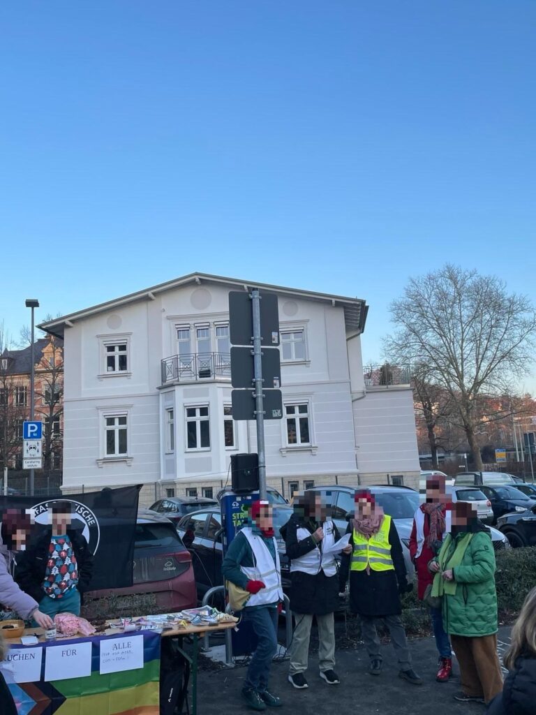 Eine Gruppe von sieben Personen steht im Freien vor einem dreistöckigen weißen Gebäude mit grauen Akzenten. Einige Personen tragen Warnwesten und ihre Gesichter sind unkenntlich gemacht. Im Vordergrund ist ein Tisch aufgebaut, auf dem Speisen und Informationsmaterialien liegen. Ein Regenbogenbanner mit der Aufschrift "Kuchen für alle (für 0€ gegen...) [unklar]" ist an der Vorderseite des Tisches befestigt. Hinter dem Tisch stehen die Personen, einige von ihnen scheinen zu sprechen oder zu gestikulieren. Im Hintergrund sind geparkte Autos und ein klarer blauer Himmel sichtbar. Am linken Bildrand ist ein Verkehrsschild zu sehen.