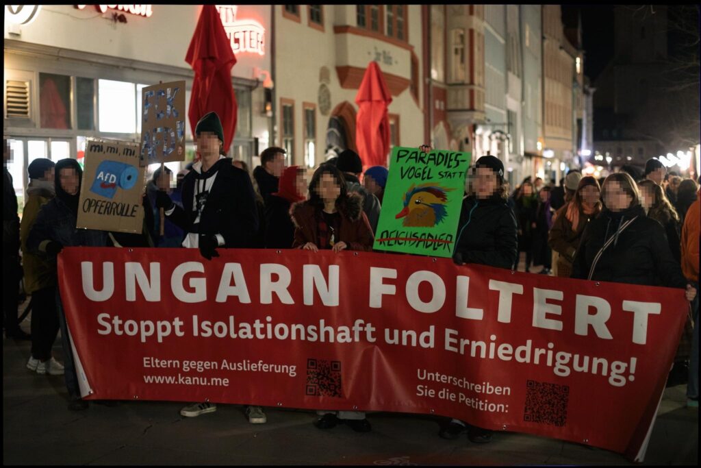 Eine nächtliche Demonstration in einer Altstadt. Eine Gruppe von Menschen hält ein großes rotes Banner mit der Aufschrift "UNGARN FOLTERT - Stoppt Isolationshaft und Erniedrigung!". Darunter steht "Eltern gegen Auslieferung" sowie eine Webseite und ein Aufruf, eine Petition zu unterschreiben, mit QR-Codes (https://weact.campact.de/petitions/keine-auslieferungen-nach-ungarn-maja-muss-zuruckkommen). Einige Demonstranten halten zusätzliche Schilder, darunter eines mit "PARADIESVOGEL statt REICHSADLER" auf grünem Hintergrund und ein anderes mit "mi, mi, mi, AfD Opferrolle". Im Hintergrund sind historische Gebäude mit roten Sonnenschirmen zu sehen. Die Straße ist voller Menschen.